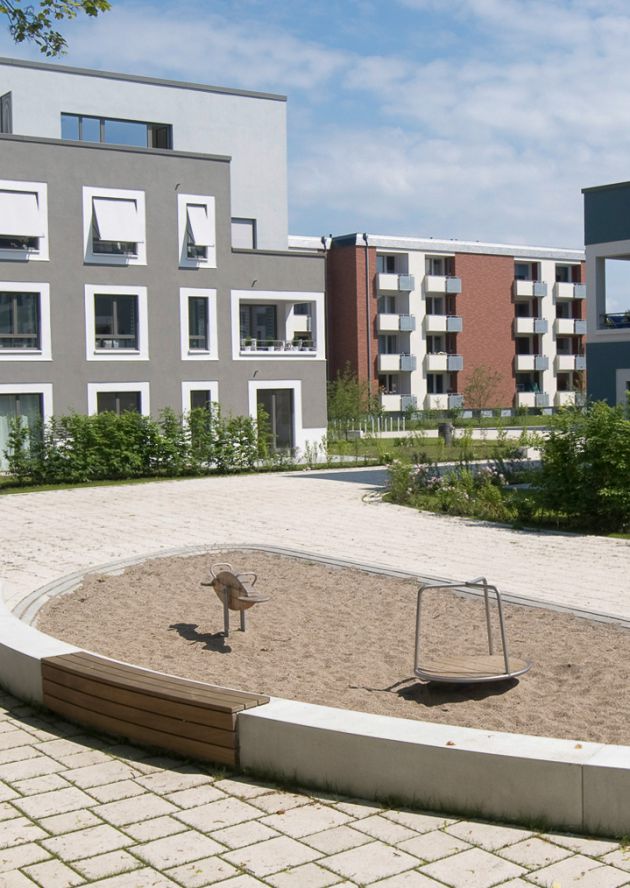 Umringt von Wohngebäuden befinden sich im Vordergrund Spielelemente in dem Sandbecken, dessen Begrenzung in Kombination mit Holzelementen eine Sitzgelegenheit bildet