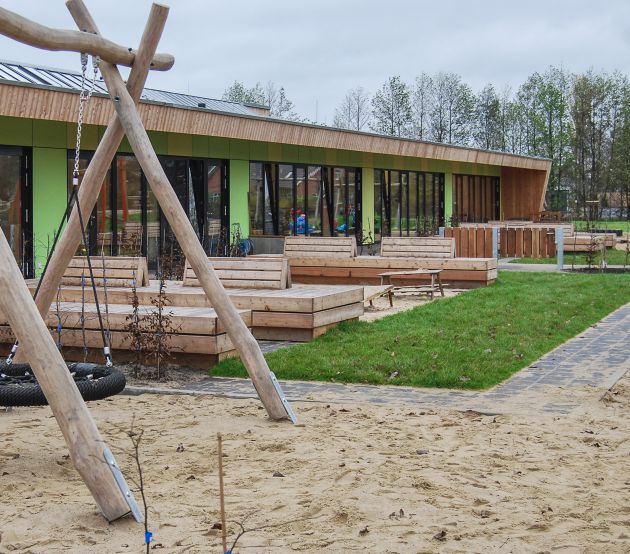 Blick auf das Gebäude mit großer Reifenschaukel auf Sandfläche im Vordergrund, neben dem Spielplatz befindet sich eine Grünfläche mit hölzernen Sitzbänken