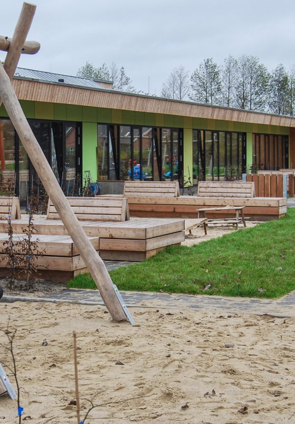 Blick auf das Gebäude mit großer Reifenschaukel auf Sandfläche im Vordergrund, neben dem Spielplatz befindet sich eine Grünfläche mit hölzernen Sitzbänken